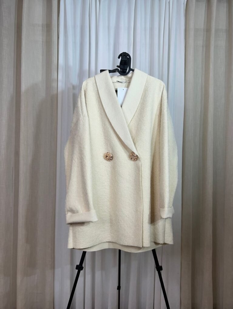 Manteau oversize crème avec boutons dorés suspendu sur un cintre — Mesdames Akbelles