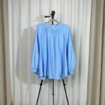 Chemise ample et fluide couleur bleu ciel sur cintre avec col froncé, boutons avant et manches longues bouffantes.
