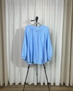 Chemise ample et fluide couleur bleu ciel sur cintre avec col froncé, boutons avant et manches longues bouffantes.