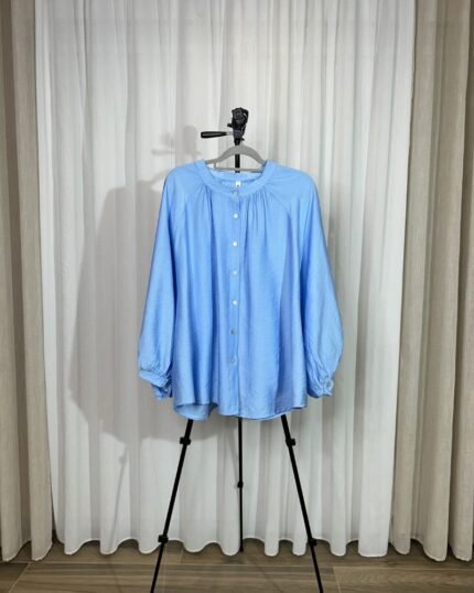 Chemise ample et fluide couleur bleu ciel sur cintre avec col froncé, boutons avant et manches longues bouffantes.