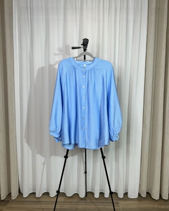 Chemise ample et fluide couleur bleu ciel sur cintre avec col froncé, boutons avant et manches longues bouffantes.