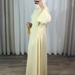 Robe longue style chemise couleur jaune pastel avec manches évasées, portée de profil avec un hijab blanc.