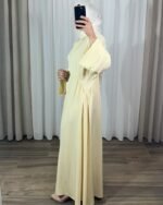 Robe longue style chemise couleur jaune pastel avec manches évasées, portée de profil avec un hijab blanc.