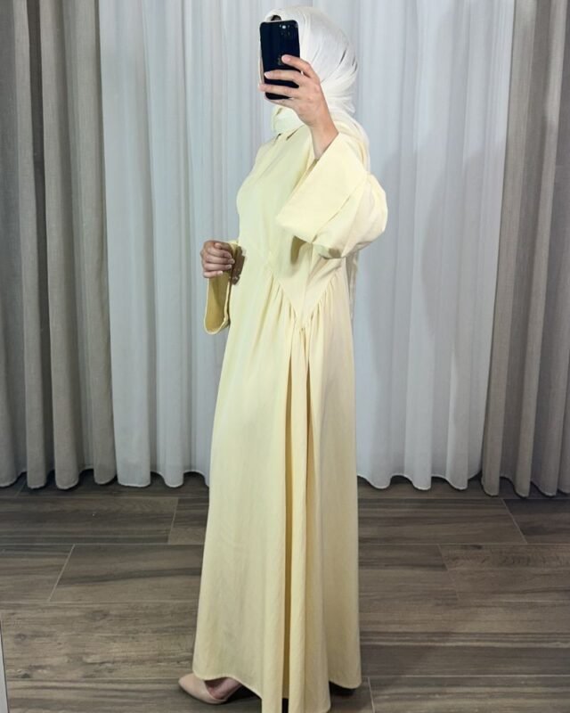 Robe longue style chemise couleur jaune pastel avec manches évasées, portée de profil avec un hijab blanc.
