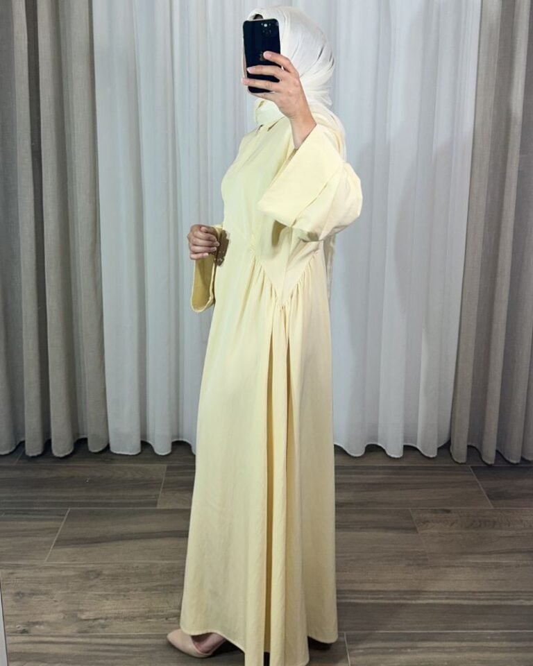 Robe longue style chemise couleur jaune pastel avec manches évasées, portée de profil avec un hijab blanc.